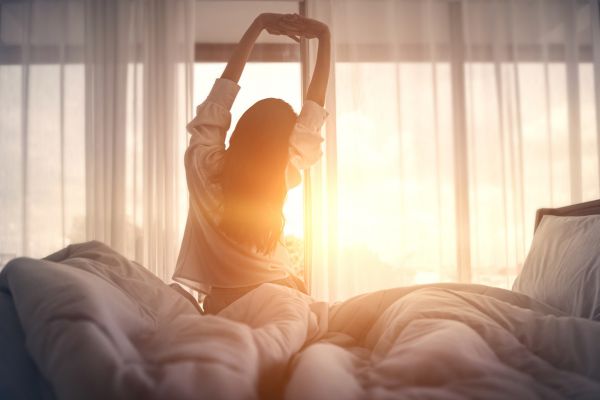 A woman sits on a bed stretching her arms overhead as warm morning sunlight streams through sheer curtains, symbolizing refreshed sleep and a natural wake-up routine.