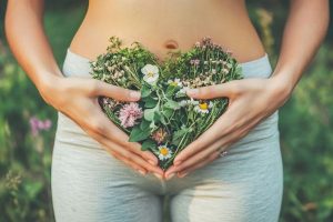Hands gently hold a heart-shaped arrangement of wildflowers over a woman’s lower abdomen, symbolizing feminine health, fertility, and holistic women’s wellness.