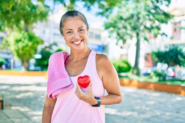 Smiling woman outdoors in workout attire holding a red heart-shaped object, with a towel over her shoulder and greenery in the background.