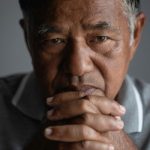Close-up portrait of an older man resting his clasped hands near his face, looking thoughtfully into the camera with a serious, reflective expression. The image features soft lighting and a neutral background, conveying themes of emotional struggle, contemplation, and men's mental health.