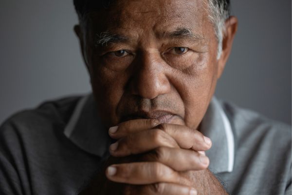 Close-up portrait of an older man resting his clasped hands near his face, looking thoughtfully into the camera with a serious, reflective expression. The image features soft lighting and a neutral background, conveying themes of emotional struggle, contemplation, and men's mental health.