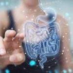 Close-up of a person reaching toward a digital holographic illustration of the digestive system, including the stomach and intestines, surrounded by interconnected data points and network lines. The image represents gut health, the microbiome, digestive wellness, and the connection between technology and human health.