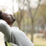 A man sits on a park bench with his hands clasped behind his head and his eyes closed, leaning back in a relaxed posture. He wears a light gray sweater, and the softly blurred background shows trees and a pathway, creating a calm, peaceful outdoor setting.