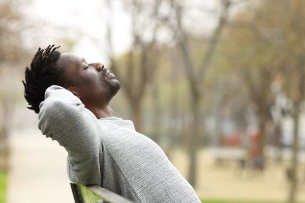 A man sits on a park bench with his hands clasped behind his head and his eyes closed, leaning back in a relaxed posture. He wears a light gray sweater, and the softly blurred background shows trees and a pathway, creating a calm, peaceful outdoor setting.